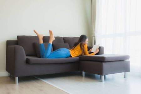 Portrait young asian woman read book on sofa chair with pillow in living room interiorの写真素材