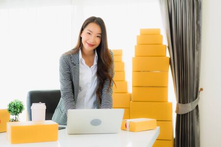 Portrait beautiful young asian business woman work from home with laptop mobile phone with cardboard box ready for shipping online shoppingの写真素材