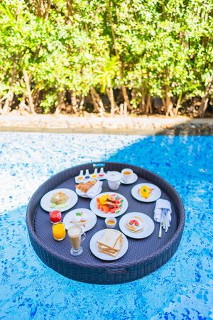 Floating breakfast set in tray with fried egg omelette sausage ham bread fruit milk juice coffee and other around swimming poolの写真素材