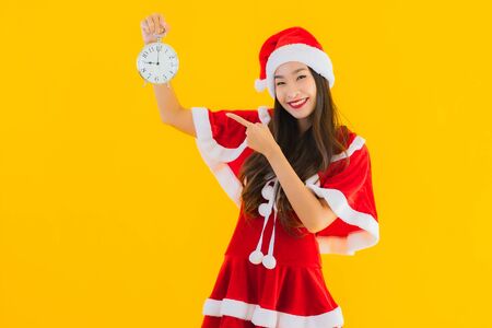 Portrait beautiful young asian woman wear christmas clothes and hat show clock or alarm on yellow isolated backgroundの写真素材