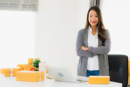 Portrait beautiful young asian business woman work from home with laptop mobile phone with cardboard box ready for shipping online shoppingの写真素材