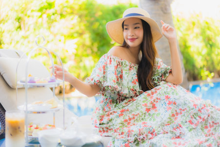 Beautiful portrait young asian woman with afternnoon tea set with coffee sit on chair around swimming pool in hotel resortの写真素材