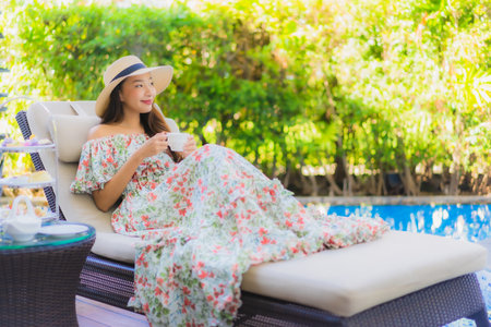 Beautiful portrait young asian woman with afternnoon tea set with coffee sit on chair around swimming pool in hotel resortの写真素材