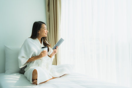 Young asian woman with coffee cup and read book on bed in bedroom interiorの写真素材