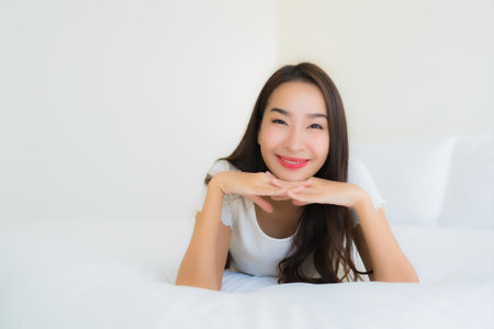 Portrait beautiful young asian woman relax happy smile on bed with white pillow blanket interior of bedroomの写真素材