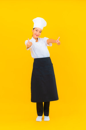 Portrait beautiful young asian woman in chef or cook uniform with hat on yellow isolated backgroundの写真素材
