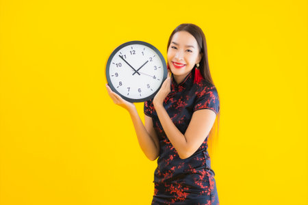 Portrait beautiful young asian woman wear chinese dress show clock or alarm on yellow isolated backgroundの写真素材
