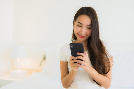 Portrait beautiful young asian woman use smart mobile phone on bed with white pillow blanket in bedroom interiorの写真素材