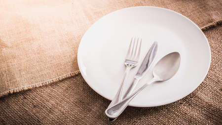 Empty white plate spoon, fork, knife on sackcloth background