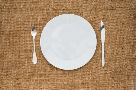 Empty white plate   fork, knife on sackcloth background