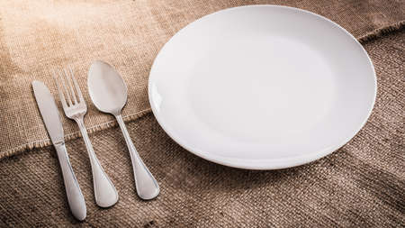 Empty white plate spoon, fork, knife on sackcloth backgroundの写真素材
