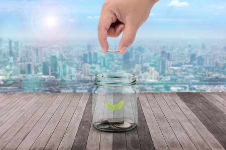 Male hand putting money coins in clear bottle and little tree on wooden Plank and green nature blending cityscape background concept business finance of money growth, prepare in the future or savingの写真素材