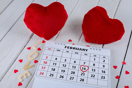 Calendar page with red hand written heart highlight on February 14 of Saint Valentines day with red Heart shape and Wooden letters word "LOVE" on white wooden tableの写真素材