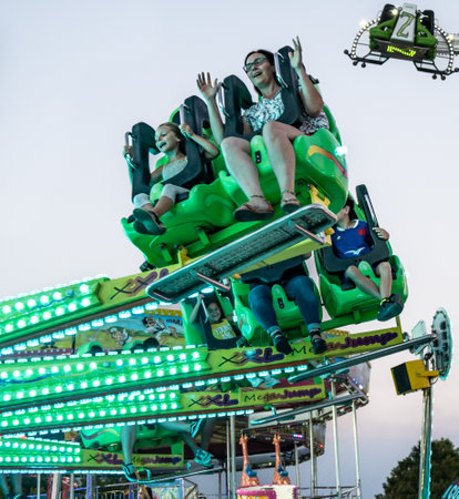 Group of people laughing and enjoying a funfair ride at full speed in an amusement park at sunset full of color and attractions.のeditorial素材