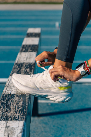 Hands of a young female runner tying the laces of her running shoes to jump an obstacle on a blue running track on a sunny day.の写真素材