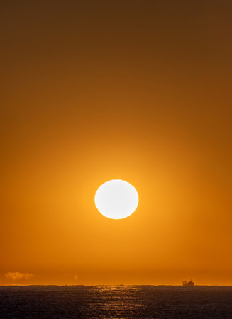 Orange sky with a bright yellow dawn sun illuminating a fishing boat sailing through the Mediterranean Sea and the first rays of the dawn sun creating optical illusions and mirages on the horizon.の写真素材