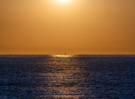 First lights of dawn backlit and illuminating the silhouette of a fishing boat sailing through the calm blue waters of the Mediterranean Sea with the brightness of the sun reflected in its waters.の写真素材
