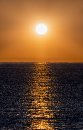 Calm sea illuminated by the orange light of the bright dawn sun illuminating the silhouettes of two groups of fishing boats fishing for PalamÃ³s shrimp in the Mediterranean Sea.の写真素材