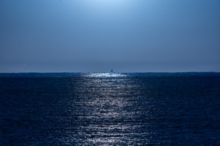 Clear, bluish night sky illuminated by moonlight with its rays of light beautifully reflected in the water of the Mediterranean Sea and a backlit fishing boat silhouette on the horizon line.の写真素材