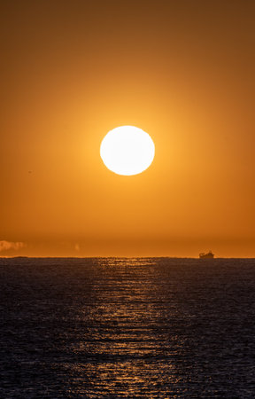 Calm sea illuminated by the orange light of the bright dawn sun and the silhouette of a fishing boat sailing through the Mediterranean Sea and various optical illusions and mirages on the horizon.の写真素材