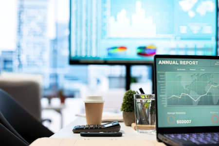 A sleek office desk setup featuring a laptop displaying an annual financial report, coffee cup, plant, and calculator in a bright workspace.