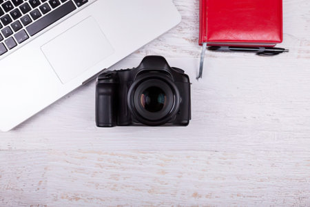 A sleek black DSLR camera placed centrally on a light wooden desk with a laptop and notebook nearby.