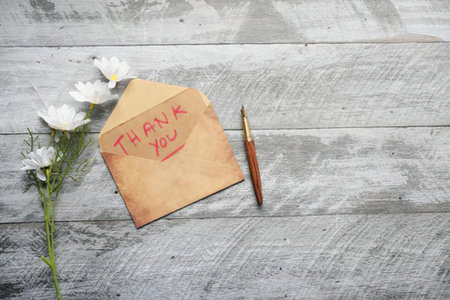 Handwritten thank you note on a brown envelope beside a white flower and pen on rustic wooden background