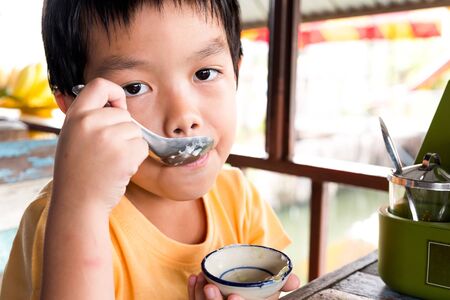 A hungry boy is sitting at the wooden table for eat dessert at restaurant with orange t-shirt.の写真素材