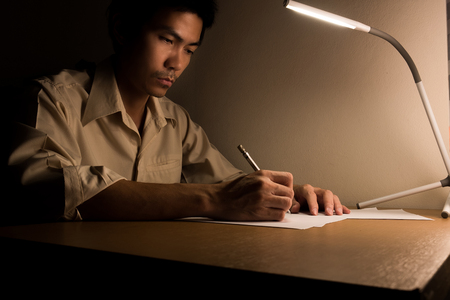A busy business man is sitting on the chair for working overtime at office at night. He use a led lamp for lighting his desk for writing and typing laptop for research information.の写真素材