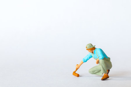 Miniature figure of a painter in a blue shirt, green overalls, and a cap, crouched while using a roller brush. on a white backgroundの写真素材