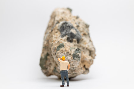 Miniature people construction worker stands next to a massive rock, scratching his head in contemplationの写真素材