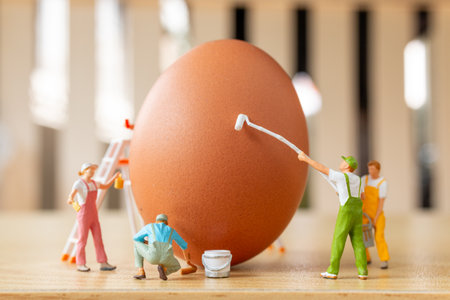 Tiny figurines painting a large brown egg. A whimsical and playful scene representing holiday preparationの写真素材