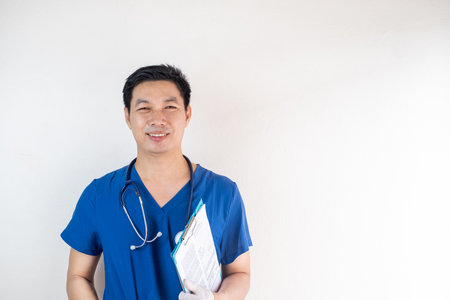 Portrait of a smiling Asian male healthcare worker wearing blue scrubs, a stethoscope, and gloves, holding a clipboardの写真素材