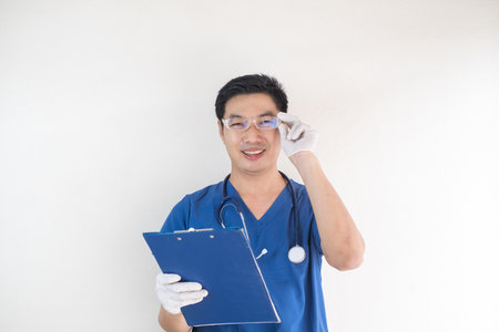Smiling Asian male nurse in blue medical scrubs, wearing eyeglasses and white gloves, holding a blue clipboardの写真素材