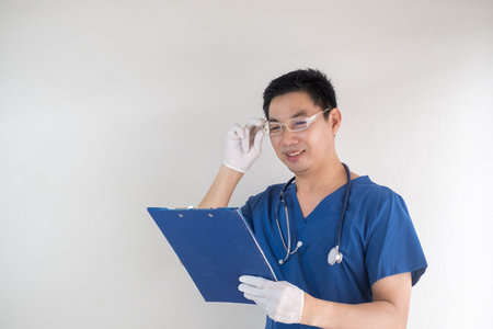 Smiling Asian male nurse in blue medical scrubs, wearing eyeglasses and white gloves, holding a blue clipboardの写真素材