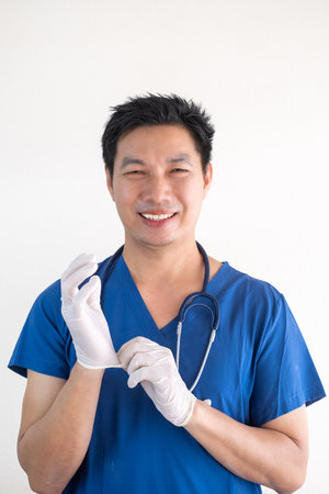 Portrait of a confident Asian male nurse wearing blue medical scrubs standing against a white wall backgroundの写真素材