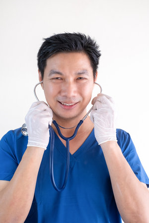 Portrait of a confident Asian male nurse wearing blue medical scrubs standing against a white wall backgroundの写真素材