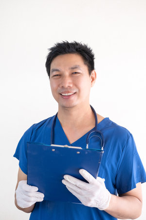 Portrait of a confident Asian male nurse wearing blue medical scrubs standing against a white wall backgroundの写真素材