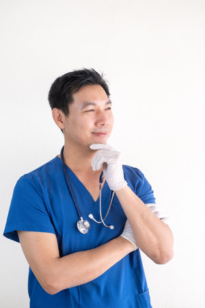 Portrait of a confident Asian male nurse wearing blue medical scrubs standing against a white wall backgroundの写真素材