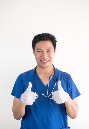 Portrait of a confident Asian male nurse wearing blue medical scrubs standing against a white wall backgroundの写真素材