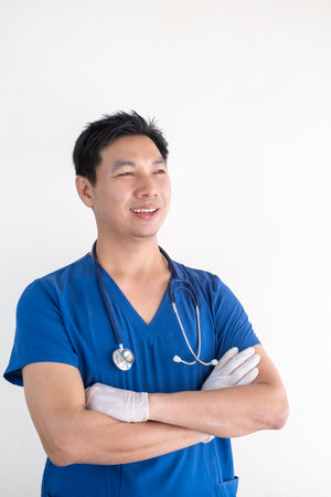 Portrait of a confident Asian male nurse wearing blue medical scrubs standing against a white wall backgroundの写真素材