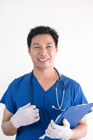 Portrait of a confident Asian male nurse wearing blue medical scrubs standing against a white wall backgroundの写真素材