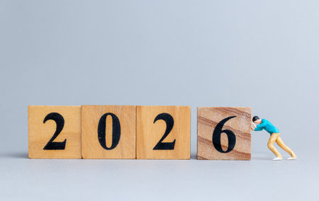 Miniature figure pushes wooden block to reveal 2026, symbolizing transition, progress, and...