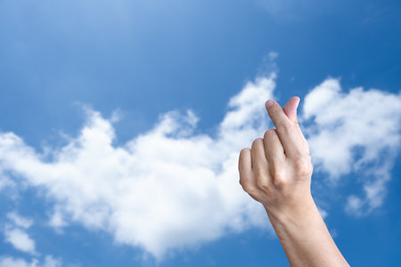 A close-up hand showing a Korean finger heart gesture against a bright blue sky with clouds.の写真素材