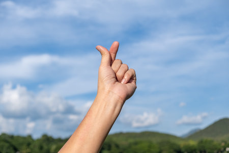 A human hand showing a Korean finger heart gesture against a natural outdoor background with blue sky and cloudsの写真素材