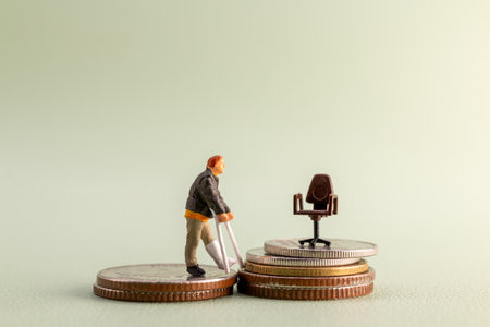 Conceptual photo of a miniature man with crutches standing on coins, looking at an office chair placed on higher stacks of coins, symbolizing challenges, inequality, and career obstaclesの写真素材