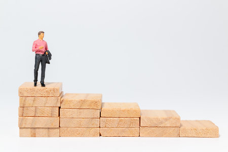 Miniature businessman standing on top of rising wooden blocks, symbolizing achievement, career growth, and reaching goalsの写真素材