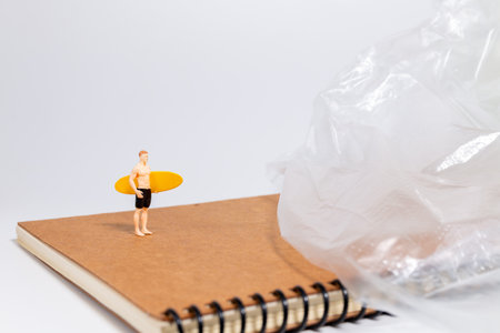 Tiny surfer figure stands on a notebook beside a sheet of plastic, symbolizing environmental issues, pollution, and ocean awareness in a creative concept scene.の写真素材