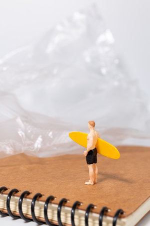 Tiny surfer figure stands on a notebook beside a sheet of plastic, symbolizing environmental issues, pollution, and ocean awareness in a creative concept scene.の写真素材