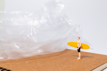 Tiny surfer figure stands on a notebook beside a sheet of plastic, symbolizing environmental issues, pollution, and ocean awareness in a creative concept scene.の写真素材